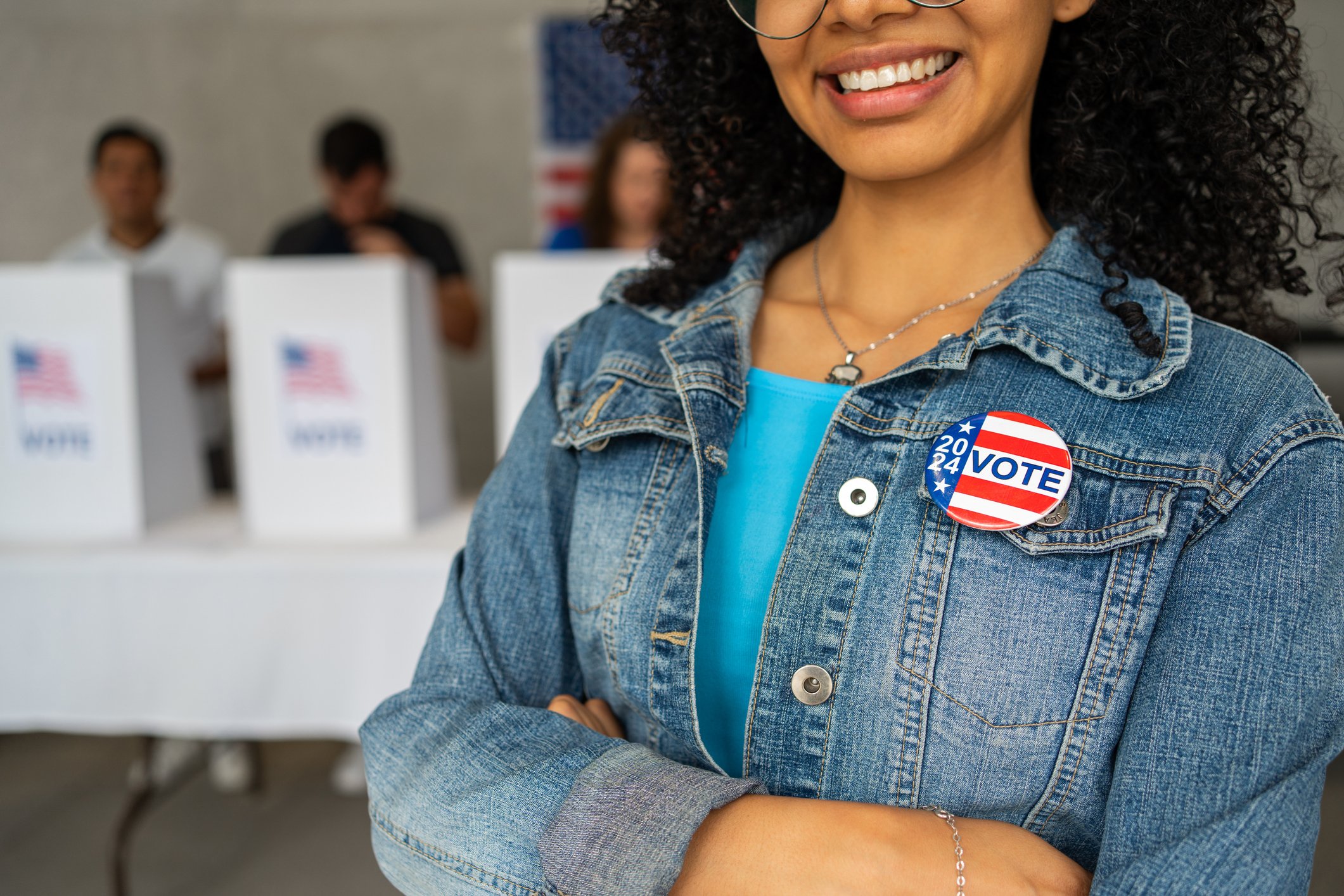 close up on a 2024 vote pin button of a young African American woman smiling at a US election polling station. In the background a diverse group of people votes in voting booths, democracy in America close up on a 2024 vote pin button of a young African American woman smiling at a US election polling station. In the background a diverse group of people votes in voting booths, democracy in America